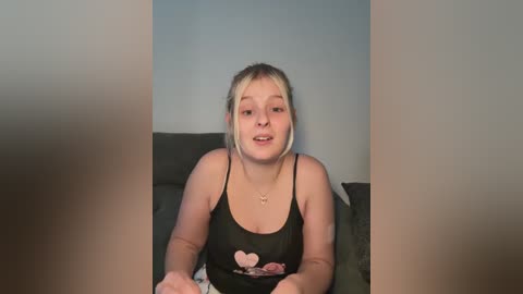 A video of a young Caucasian woman with blonde hair, wearing a black spaghetti-strap tank top with pink heart design, sitting on a couch against a plain, light blue wall.