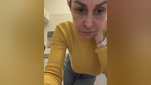 Media: Video of a light-skinned woman with short brown hair, wearing a mustard yellow ribbed sweater and blue jeans, leaning forward, looking serious, in a modern kitchen with white tiles and stainless steel appliances.