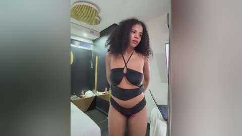 Video of a curvy, light-skinned woman with curly hair, wearing black lingerie, standing in a modern bedroom with black walls, white bed, and circular lighting.