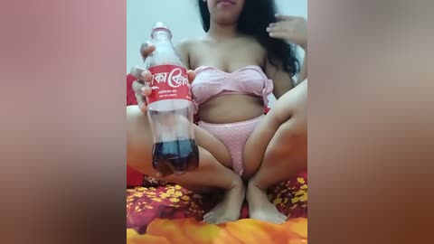 Video of an Asian woman with long black hair, squatting on a red and yellow patterned bedspread, wearing pink lingerie, holding a Coca-Cola bottle.