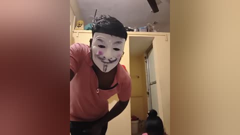 Video of a person wearing a creepy clown mask, leaning forward, with a red shirt and black pants, in a dimly lit hallway with yellow walls and a ceiling fan.