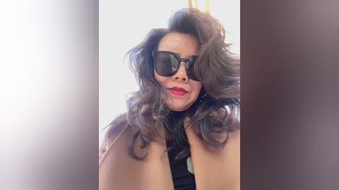 Video of a smiling woman with wavy brown hair, large black sunglasses, and a tan jacket, set against a bright, blurred background.