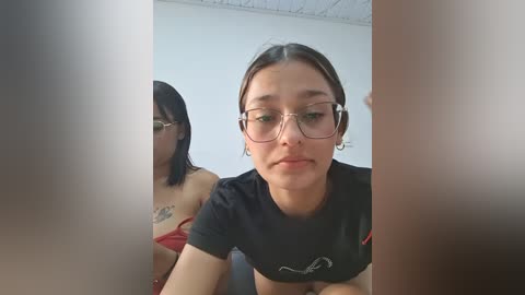 Video of a young woman with glasses, dark hair, and a black t-shirt, sitting indoors; partially obscured by a transparent curtain.