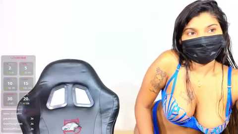 Video of a Latina woman with long black hair, wearing a blue bikini top, black face mask, and a Los Angeles Dodgers baseball jersey. She sits on a black gaming chair, against a white background.