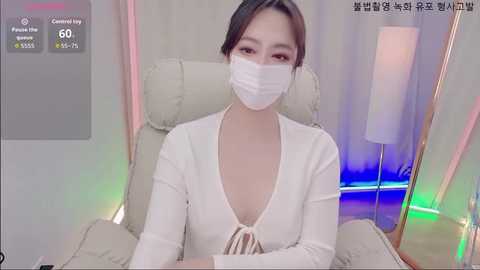Media: A video of an East Asian woman with light skin, dark hair, wearing a white face mask and a low-cut white top, seated in a beige armchair with a glowing blue light background.
