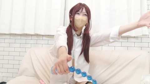 Video of a young Asian woman with long, red hair, wearing a white mask and a white top, reaching out with a blue-bead necklace in a tiled bathroom.