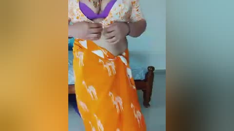 Video of a light-skinned woman in a purple bra, lifting her shirt to reveal a small bust, wearing orange pants with a white pattern. She sits on a bed with a wooden headboard, in a dimly lit room.