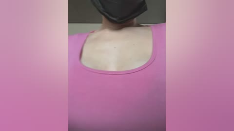 A video of a person's upper torso, wearing a pink tank top and a black face mask. The background is blurred and light-colored, likely indoor. The focus is on the chest area, with no visible facial features.