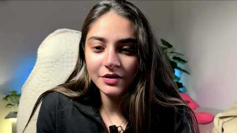 Video of a young woman with long, dark hair, wearing a black jacket, seated on a white chair in a dimly lit room with a potted plant and colorful objects in the background.