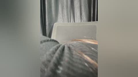 A video of a person lying face down on a bed, partially visible in a dimly lit room. The person is wearing a grey shirt and the background features a grey curtain and a white chair.