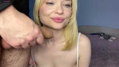 A close-up video of a blonde woman with fair skin and blue eyes, wearing a white tank top, smiling seductively while a man's hand holds his erect penis in front of her.