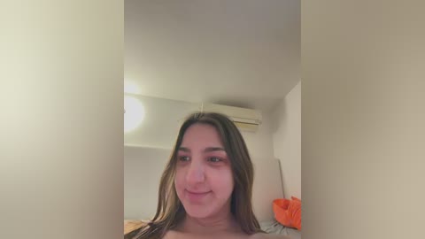 Media: A video of a young woman with light brown skin and long brown hair, smiling slightly. She is indoors, with a white ceiling fan and an orange item in the background.