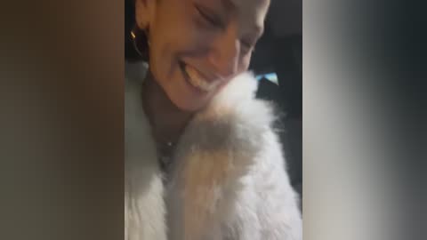 A video of a smiling young woman with short brown hair, wearing a white fluffy coat, hugging a fluffy dog. The background is dark and blurred, emphasizing the cozy moment.