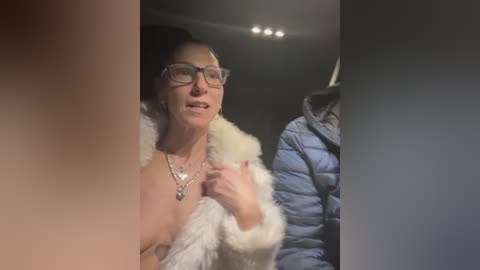 Video of a light-skinned woman with glasses, wearing a fur-trimmed jacket, and a silver necklace, sitting in a dimly lit car, with a blurred figure in the foreground.