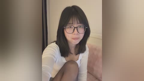 Video of an Asian woman with shoulder-length black hair, wearing glasses and a white top, sitting on a pink couch in a dimly lit room.