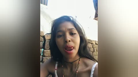 A video of a young woman with medium brown skin and long, dark hair, wearing a white tank top and multiple beaded necklaces, set against a rustic, stone wall background. She has a neutral expression, with slightly parted lips and her tongue sticking out.