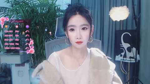 Video of an East Asian woman with fair skin and dark hair, wearing a beige coat and pearl necklace, seated in a room with a plant, computer monitor, and a studio light.
