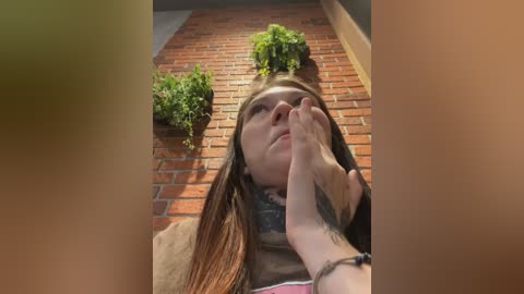 A video of a young woman with long brown hair, wearing a brown shirt, lying on a brick patio with green potted plants. Her hand is touching her face, revealing a tattoo on her wrist.