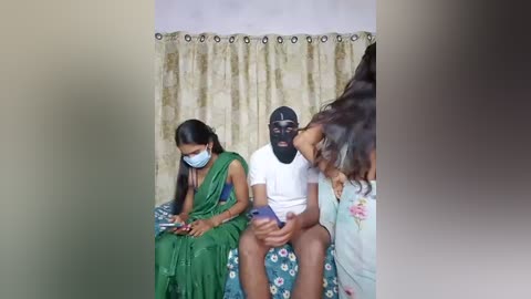 Video of three people sitting on a bed in a dimly lit room with beige curtains. A man in a black mask, white shirt, and shorts, a woman in a green sari, and a child in a floral dress.