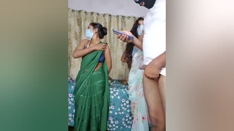 Video of three women in a small room with floral curtains, one in a green sari and mask, another in a floral dress, and a third in a white shirt, holding a phone.