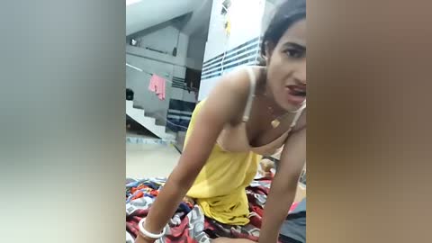 Video of a young woman with dark hair, wearing a yellow dress, leaning over a messy bed in a dimly lit room with a staircase and clothes hanging in the background.