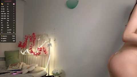 A video of a young woman with light skin and a slender physique, standing nude beside a cluttered bed with a floral lampshade and calendar.