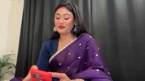A video of a South Asian woman with medium skin tone, wearing a purple sari with silver sequins, and a blue blouse. She has long, straight black hair, and is holding a red box, smiling.
