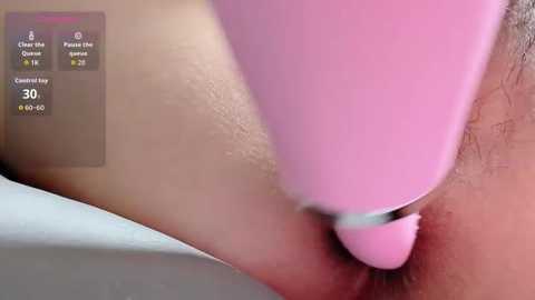 Close-up video of a pink vibrator being used on a person's vulva, with the person's pubic hair visible. The background shows a blurred setting with digital icons.