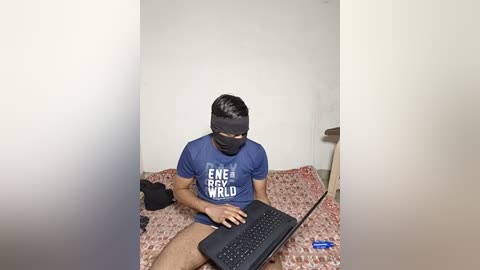 Video of a young man with short black hair, wearing a black mask and blue t-shirt, sitting on a pink and white patterned bedspread, typing on a laptop, in a minimalistic white room.