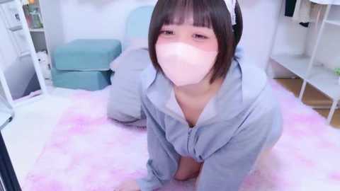 Video of an East Asian woman with short black hair, wearing a white mask, light blue hoodie, and a pink fluffy rug in a minimalist room with white furniture.