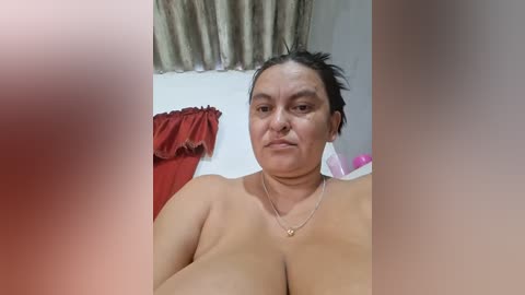 A video of a middle-aged woman with medium skin tone, dark hair, and large breasts, standing topless in a room with beige curtains, red and pink clothing items visible in the background.