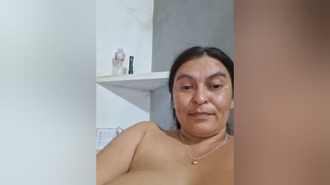 Video of a smiling, topless Latina woman with medium skin tone and shoulder-length black hair, wearing a thin necklace. She's in a bathroom with a white shelf holding toiletries and a light gray wall.