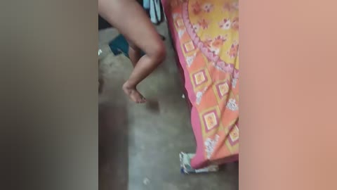 A video shows a person's bare legs, one bent, on a colorful, patterned bedspread in a dimly lit room with scattered clothes and a concrete floor.