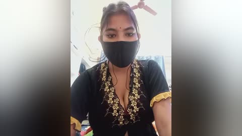 Video of a woman with medium brown skin, dark hair, and a black face mask, wearing a black blouse with gold embroidery and yellow sleeves, standing indoors with a ceiling fan visible.