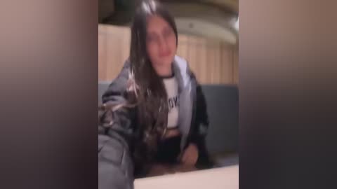 A blurry video shows a person with long, dark hair in a black jacket and white shirt, seated in a vehicle with beige upholstery.