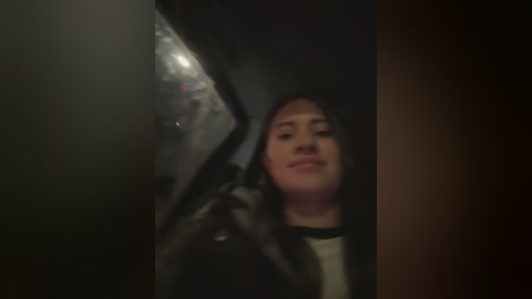 A dimly lit video captures a young woman with light skin and dark hair, smiling while holding a black and silver smartphone in front of her face.