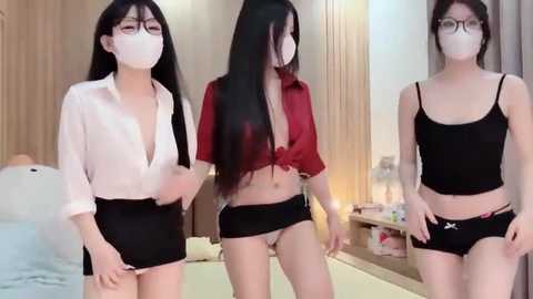 Video of three East Asian women in lingerie, face masks, and glasses, in a modern bedroom setting with wooden furniture and a bed.