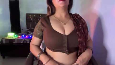 A video of a South Asian woman with medium skin tone and dark brown hair, wearing a brown, low-cut blouse with lace detailing, a maroon saree, and gold jewelry. She sits in a modern bedroom with a colorful TV and a bed in the background.