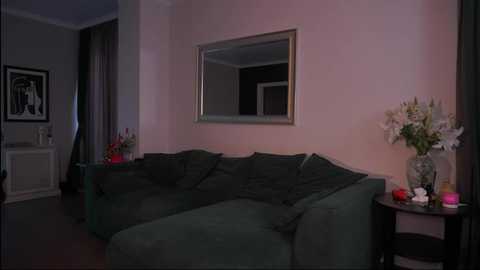 A dimly lit living room with a plush green sectional sofa, a large rectangular mirror, and two black side tables with floral arrangements and a red candle.