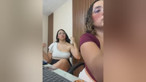 Video of two women in an office. One woman, with glasses, sits at a desk, wearing a white one-shoulder top and black skirt, looking frustrated. The other, seated behind her, wears a maroon top, blushing, and appears to be watching.