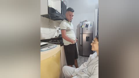 Video of a man in a white polo shirt and black shorts standing in a small, cluttered kitchen with a stainless steel fridge and a yellow washing machine. Another man in a grey shirt and glasses sits at a table.