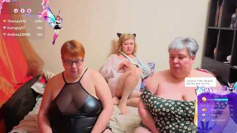 Video of three older women, one in a black leather dress, one in a floral off-shoulder top, and one in a white robe, sitting on a bed in a dimly lit room.