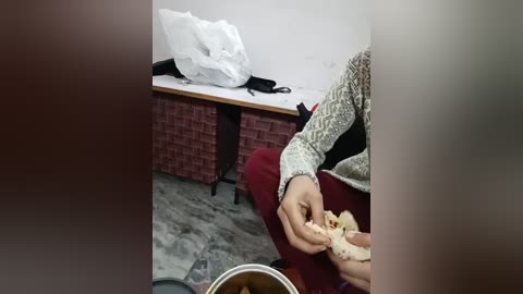 A video showing a person in a red sweater eating a sandwich, sitting on a chair in a cluttered room with a brick-patterned shelf and a messy white pillow.