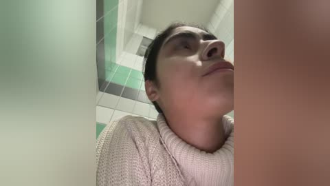 Video of a young woman with fair skin, dark hair, and a light beige sweater, captured from a low angle, inside a tiled bathroom with green and white tiles.