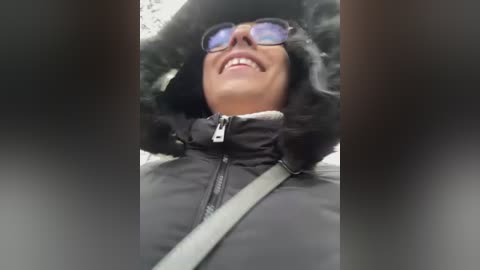 Video of a grinning person in a black winter coat, fur-lined hood, and reflective sunglasses, taken from a low angle, capturing a joyful, energetic expression.