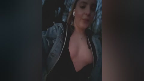 Media: A dimly lit video of a fair-skinned woman with shoulder-length hair, wearing a black top and a light jacket, smiling softly. The background is out of focus.