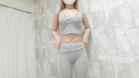 Video of a young Asian woman with fair skin, wearing a light grey, sleeveless crop top and matching leggings. She stands against a textured, white-tiled wall, with a white shower curtain on the left.