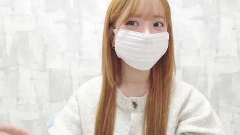 Video of a young Asian woman with long, straight, light brown hair, wearing a white surgical mask and a white, textured sweater. She gazes upward, set against a blurred, white, textured background.