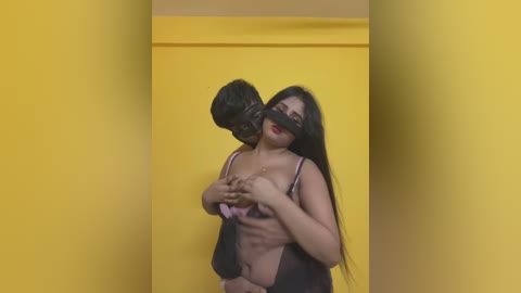 Video of two women in a BDSM scene; one is blindfolded and gagged, wearing lingerie, while the other, with long black hair, holds her. They are set against a yellow backdrop.
