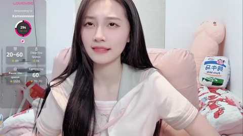 A video of a young East Asian woman with long black hair, light skin, and a slender physique, wearing a pink top and holding a plush toy, surrounded by colorful pillows in a bright, minimalist room.